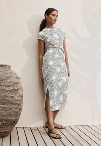 Weißes Kleid mit schwarzen geometrischen Mustern, kurzen Ärmeln, tailliertem Schnitt und seitlichem Schlitz. Getragen mit schwarzen Sandalen auf einem Holzboden neben einer geflochtenen Vase.