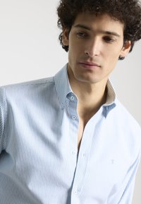 Camisa de rayas azul claro hecha de algodón, con cuello tipo button-down, un bolsillo en el pecho y un sutil logo en el lado izquierdo.