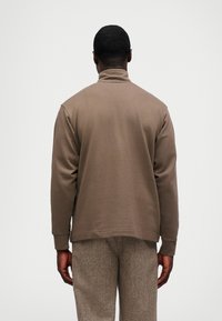 Καφέ βαμβακερή μπλούζα με ψηλό γιακά, μακριά μανίκια και χαλαρή εφαρμογή, συνδυασμένη με υφασμάτινο παντελόνι σε μπεζ απόχρωση.