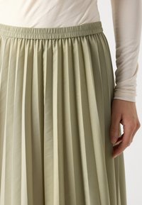 Plisserte, lys grønn skjørt laget av glatt stoff med elastisk midje; nærbilde viser tekstur og draperingsdetaljer.
