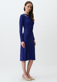Langärmliges, knielanges Kleid in Dunkelblau, mit gerafften Details und Ausschnitten, kombiniert mit nudefarbenen High Heels.