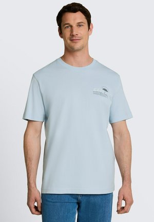 Mężczyzna ubrany w jasnoniebieski T-shirt z krótkim rękawem z małym nadrukiem tekstu i grafiki na lewej piersi, w połączeniu z niebieskimi dżinsami.