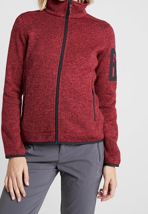 Vrouw draagt een rood fleecejack met ritssluitingen en ritstassen en grijze outdoorbroek, staat met handen nabij de heupen.