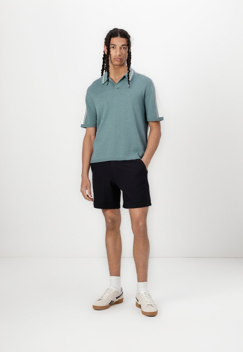 Polo à manches courtes teal avec une finition texturée et un col contrastant, associé à un short noir et des baskets légères.