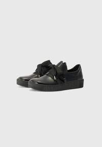 Baskets noires en cuir lisse avec une semelle en caoutchouc texturée. Elles présentent un large ruban noir noué en un nœud sur le dessus.