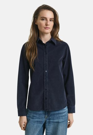 Frau mit langen braunen Haaren, die ein navyblaues, knöpfbares Cordhemd und eine blaue Jeans trägt, steht vor einem schlichten weißen Hintergrund.