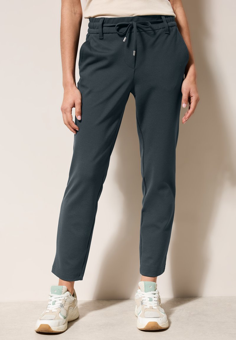 Pantalones de chándal gris oscuro con cintura elástica y cordón, con bolsillos laterales y piernas ajustadas, combinados con zapatillas de color claro.