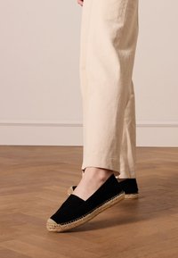 Espadrilles noires en toile avec garniture en jute naturel et semelles plates, associées à un pantalon en lin crème sur un sol en bois.