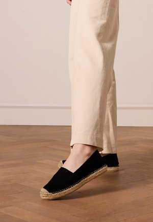 Zwarte canvas espadrilles met een natuurlijke jute rand en platte zolen, gecombineerd met crème linnen broeken op een houten vloer.