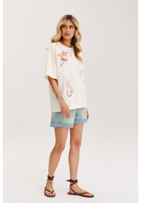 Krémové tričko s farebnými grafickými potlačami, krátke rukávy, v kombinácii s svetlými denimovými šortkami a hnedými sandálmi s ovinutými popruhmi.
