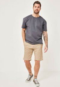 Gray cotton t-shirt, short sleeves, paired with beige shorts. Worn with gray Converse sneakers. Visible tattoos on arm and leg.