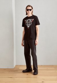 Zwarte katoenen t-shirt met een grafisch ontwerp van een leeuwenkop binnen een rode driehoek. Gecombineerd met donkere jeans en zwarte sneakers.