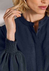 Frau, die ihre kurzen blonden Haare anpasst, trägt eine marineblaue Bluse mit gerafften elastischen Ärmelbündchen und einem geknöpften Kragen.