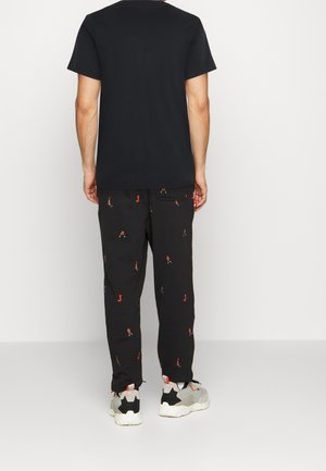 Person trägt ein einfaches schwarzes T-Shirt, schwarze Hose mit kleinen roten und weißen Mustern sowie graue und weiße Turnschuhe und steht vor einem weißen Hintergrund.