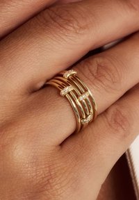 Goldene Multi-Band-Ringe mit verflochtenen Designs und kleinen weißen Edelsteinen, die die Bänder akzentuieren, vor einem strukturierten Hintergrund von Haut.