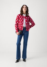 Punainen neuletakki, jossa on valkoisia sydänkuvioita, hyvin istuva malli, pyöreä kaula-aukko, yhdistetty tumma sinisiin levennettyihin farkkuihin ja mustiin kärjellisiin korkokenkiin.