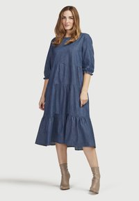 Denimmekko, jossa on kolmisen neljännestä puffi-hihkaa, kerroksellinen muotoilu ja midi-pituus. Pehmeä kangas, syvä sininen väri, yhdistettynä beigeihin nilkkureihin.