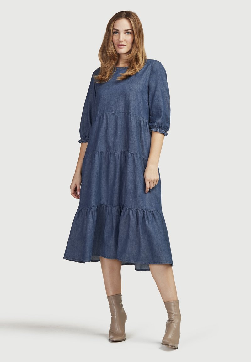 Denimmekko, jossa on kolmisen neljännestä puffi-hihkaa, kerroksellinen muotoilu ja midi-pituus. Pehmeä kangas, syvä sininen väri, yhdistettynä beigeihin nilkkureihin.