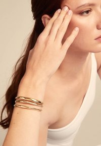 Pulseras de brazalete de metal dorado con un diseño retorcido, llevadas en una muñeca. El tono de piel es claro, destacando la textura brillante de la pulsera.