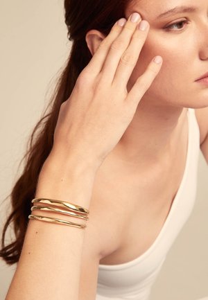 Bracciali in metallo dorato con design intrecciato, indossati su un polso. Il tono della pelle è chiaro, evidenziando la texture lucida del bracciale.