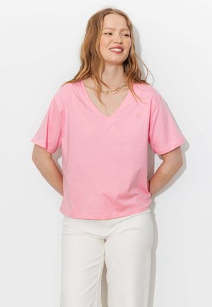 Femme souriante, portant un t-shirt rose à col en V et manches courtes et un pantalon blanc, debout les mains dans le dos sur fond blanc.