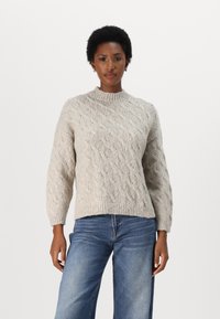 Beige strikket genser med kabelmønster, høy hals og ribbestrikkede mansjetter. Kombinert med blå denimjeans med en avslappet passform.