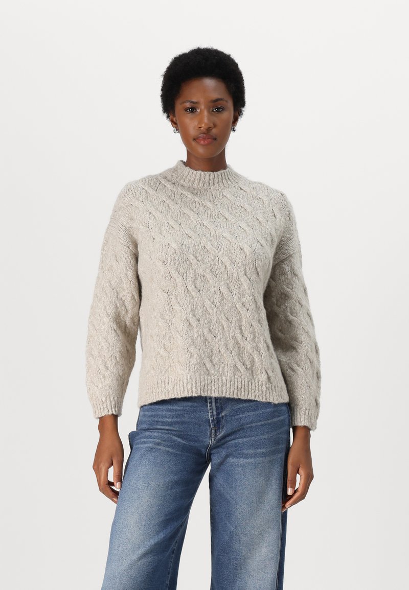 Beige strikket genser med kabelmønster, høy hals og ribbestrikkede mansjetter. Kombinert med blå denimjeans med en avslappet passform.