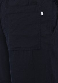Pantalon bleu marine en tissu léger, doté d'une poche plaquée et d'une discrète étiquette de marque sur le côté. Texture lisse, design décontracté.