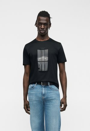 Giovane uomo con pelle scura che indossa una t-shirt nera con una grafica di un'auto e jeans beige sbiaditi, in piedi davanti a uno sfondo bianco semplice.