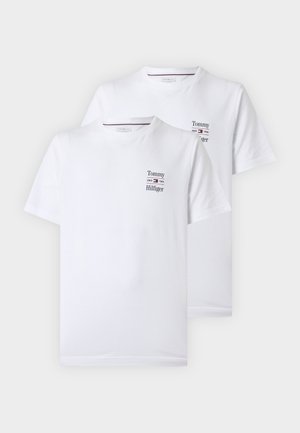 Mand stående med hænderne i lommerne iført hvid Tommy Hilfiger T-shirt, løstsiddende lyseblå jeans og mørkebrune sko på en ensfarvet baggrund.