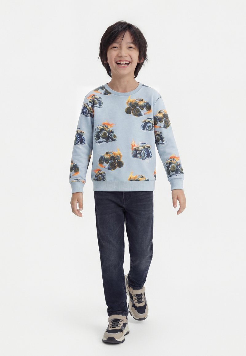 Χαμογελαστό αγόρι που περπατά, φοράει ανοιχτό μπλε φούτερ με στάμπες φλεγόμενων φορτηγών τέρας, σκούρο τζιν και μπεζ και μαύρα αθλητικά παπούτσια.