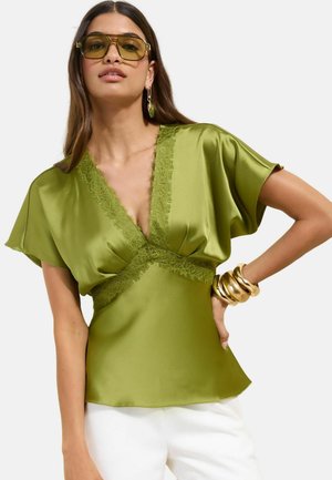Kobieta ubrana w oliwkowo-zieloną satynową bluzkę z koronkową lamówką, białe spodnie, duże złote bransolety, zielone wiszące kolczyki i duże żółte okulary przeciwsłoneczne.