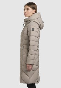 Podloženi plašč do kolen v svetlo bež barvi z diagonalen vzorcem, prilegajočo kapuco in črnimi akcenti na rami ter zadrgi za zapiranje.