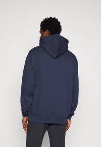Sweat-shirt à capuche bleu marine en tissu doux, avec une coupe décontractée et des poignets larges. La vue arrière montre les détails de la capuche et de l'ourlet.