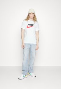 T-shirt in cotone bianco con logo "nike" nero e swoosh rosso. Jeans azzurri chiari, sneaker bianche con accenti verdi e gialli. Cappellino beige chiaro.