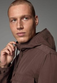 Chaqueta ligera marrón con capucha, con cierre de cremallera y cordón ajustable. Textura suave y detalles de costura visibles.
