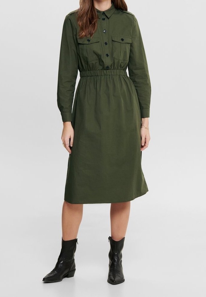 Femme portant une robe midi boutonnée à manches longues couleur olive avec taille élastique, poches poitrine, et bottines noires, sur un fond uni.