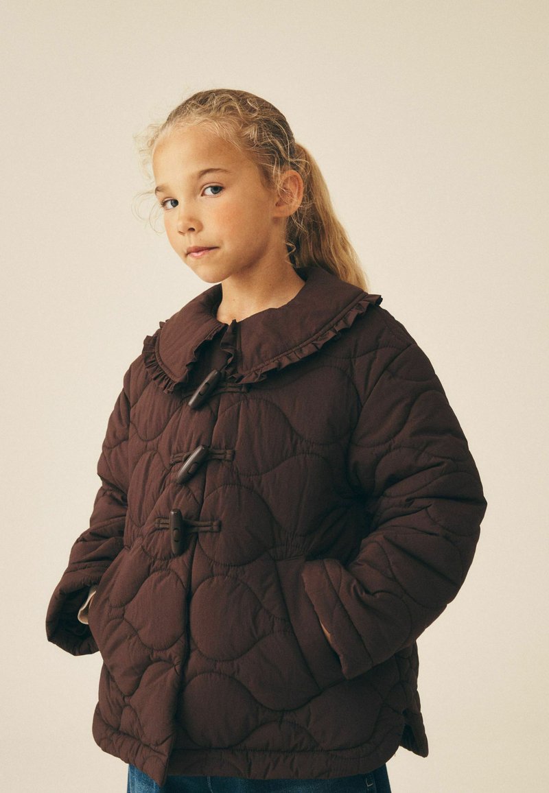 Jeune fille aux cheveux blonds attachés portant une veste matelassée marron foncé avec des boutons à bascule et les mains dans les poches, sur fond uni.