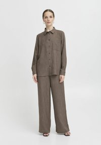 Chemise boutonnée marron texturée avec une poche poitrine unique, associée à un pantalon large marron. Les deux vêtements ont une coupe décontractée et un design minimaliste.