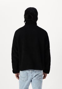 Schwarze Fleecejacke mit hohem Kragen, strukturiertem Stoff und lockerer Passform. Kombiniert mit hellblauen Jeans aus Denim mit gestickten Gesäßtaschen.