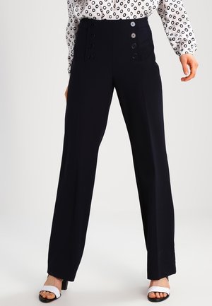 Schwarz hochtaillierte, weit geschnittene Hosen mit doppelten Knopfreihen auf der Vorderseite, kombiniert mit einer weißen Bluse mit schwarzen Kreis-Mustern und weißen offenen Sandalen mit Absatz.