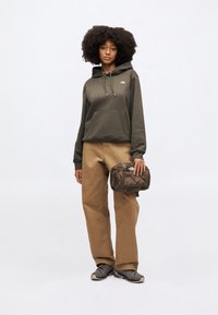 Olivovo zelená mikina s kapucňou a predným vreckom, v kombinácii s béžovými širokými nohavicami a tmavou obuvou. Drží hnedú textúrovanú tašku s logom.