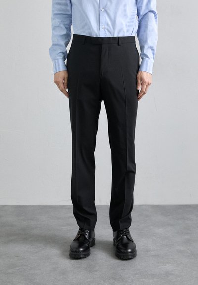 Pantaloni neri sartoriali con frontale piatto, vestibilità slim e tessuto liscio, abbinati a scarpe in pelle nera, su uno sfondo grigio chiaro.