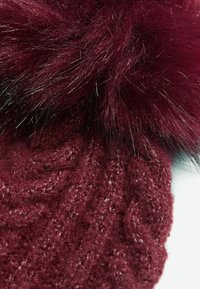Căciulă tricotată burgundy cu un model texturat, având un pom-pom pufos din blană artificială în vârf. Materialele moi și călduroase oferă un aspect confortabil.