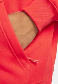 Korallfärgad sweatshirt med känguruficka, ribbade ärmslut och snörningsdetalj med metalltips. Slät textur med en figurnära design.