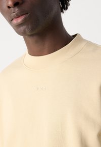 Gros plan d'une personne portant un sweat-shirt beige avec un col côtelé et le mot HUGO embossé sur la poitrine.