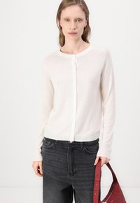 Hvit lett cardigan med lange ermer og knappestolpe, kombinert med mørkegrå jeans og en rød veske. Myk tekstur, enkelt design.