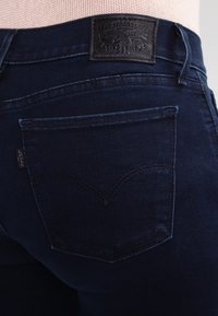 Mörkblå denimjeans med en slät textur, med en brun läderpatch på baksidan och en klassisk fickdesign med sömnadsdetaljer.