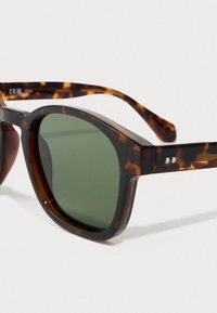 Kivimäiseksi acetaatiksi valmistetut aurinkolasit, joissa on paksut neliömäiset kehykset ja tummanvihreät linssit, levättävät valkoisella pinnalla.