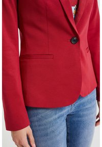 Röd blazer med en enda svart knapp, slät tyg, skräddarsydd design, två framfickor, ihopparad med blå denimjeans.